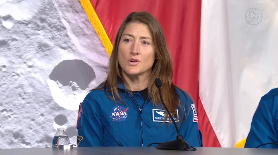  A woman with long dark hair wears a blue flight suit with the circular NASA logo on it and sits behind a desk with a microphone. 