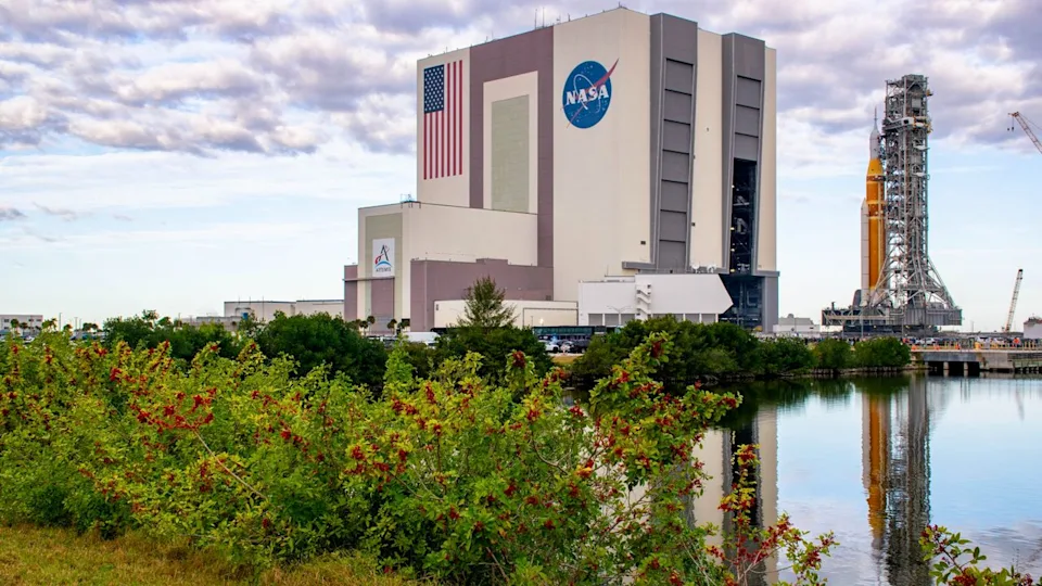  An orange rocket sits on a mobile scaffold as it rolls out of a large white building with the American flag and NASA meatball logo on the side. 