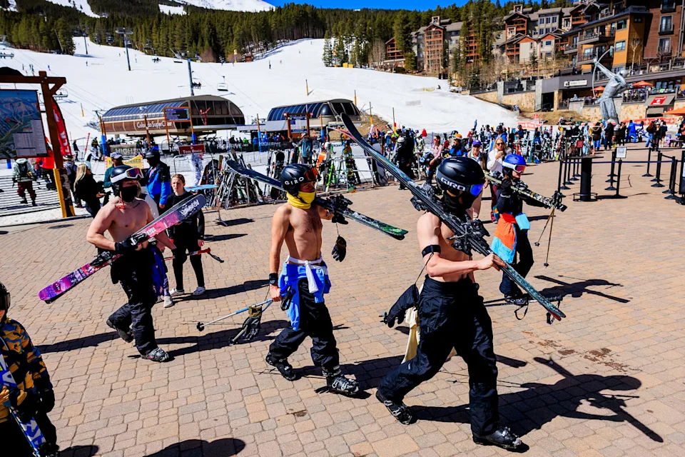 Three shirtless men wearing helmets and goggles carry ski equipment across a stone walkway at a ski resort. (Michael Ciaglo / Getty Images)