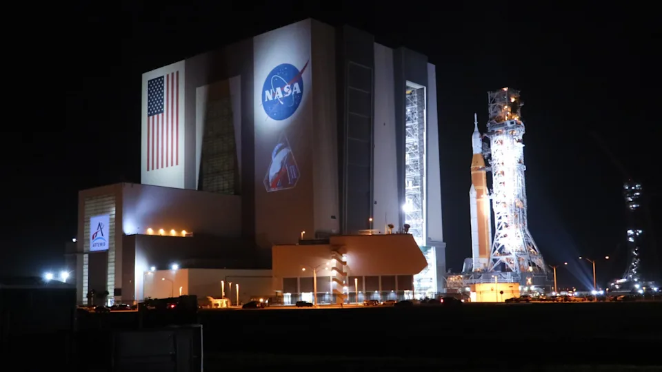 a white-and-orange rocket rolls out of a large hangar at night 