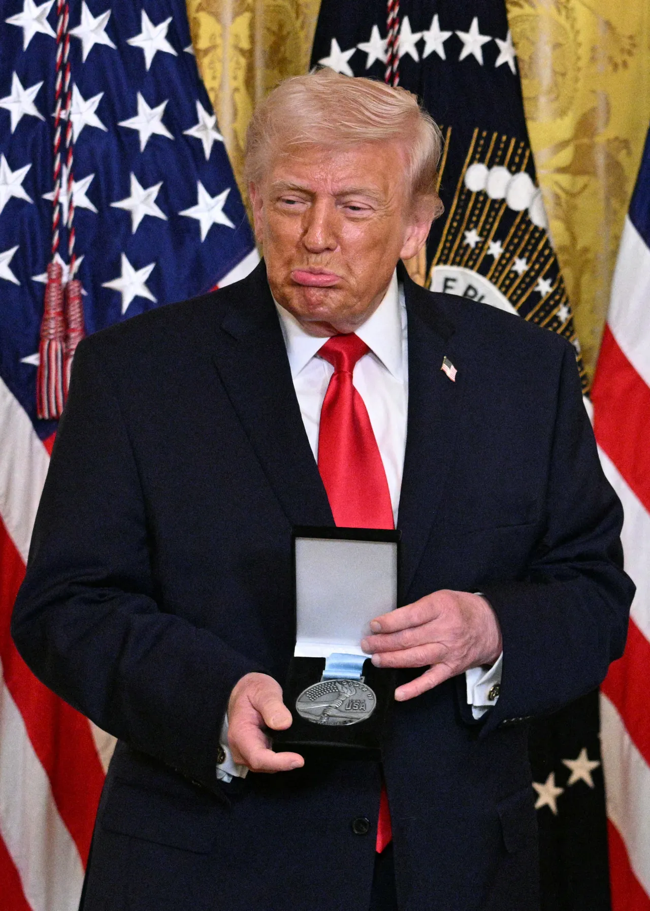 US President Donald Trump reacts after US bobsledder Kaillie Armbruster Humphries (out of frame) awarded him with an Ikkos award during a Women's History Month event in the East Room of the White House in Washington, DC, on March 12, 2026. Medal-winning team USA athletes can award an Ikkos award to recognize people who have been supportive in their journey to the Games. (Photo by Jim WATSON / AFP via Getty Images)