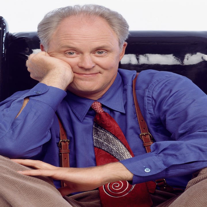 Person sitting on a chair, wearing a blue shirt and patterned tie with suspenders, smiling with chin resting on hand