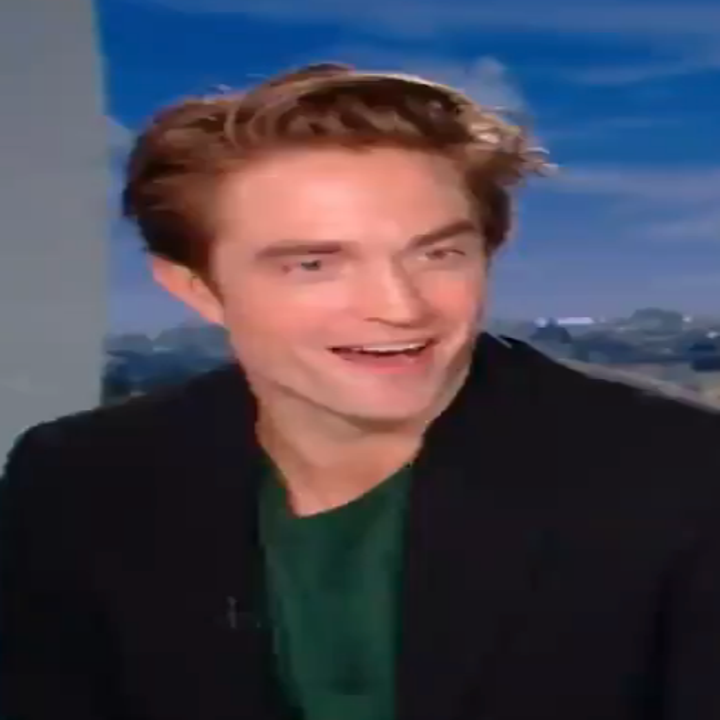 A person with a green shirt and black blazer sits smiling in a studio setting, with a cityscape in the background