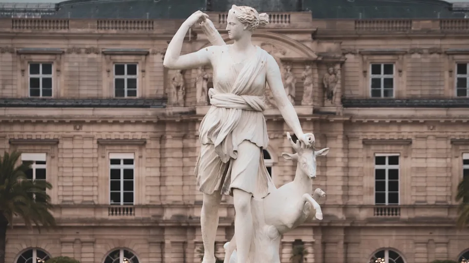 a white statue of a female figure in a toga holding a quiver of arrows, placing her hand on the head of a deer