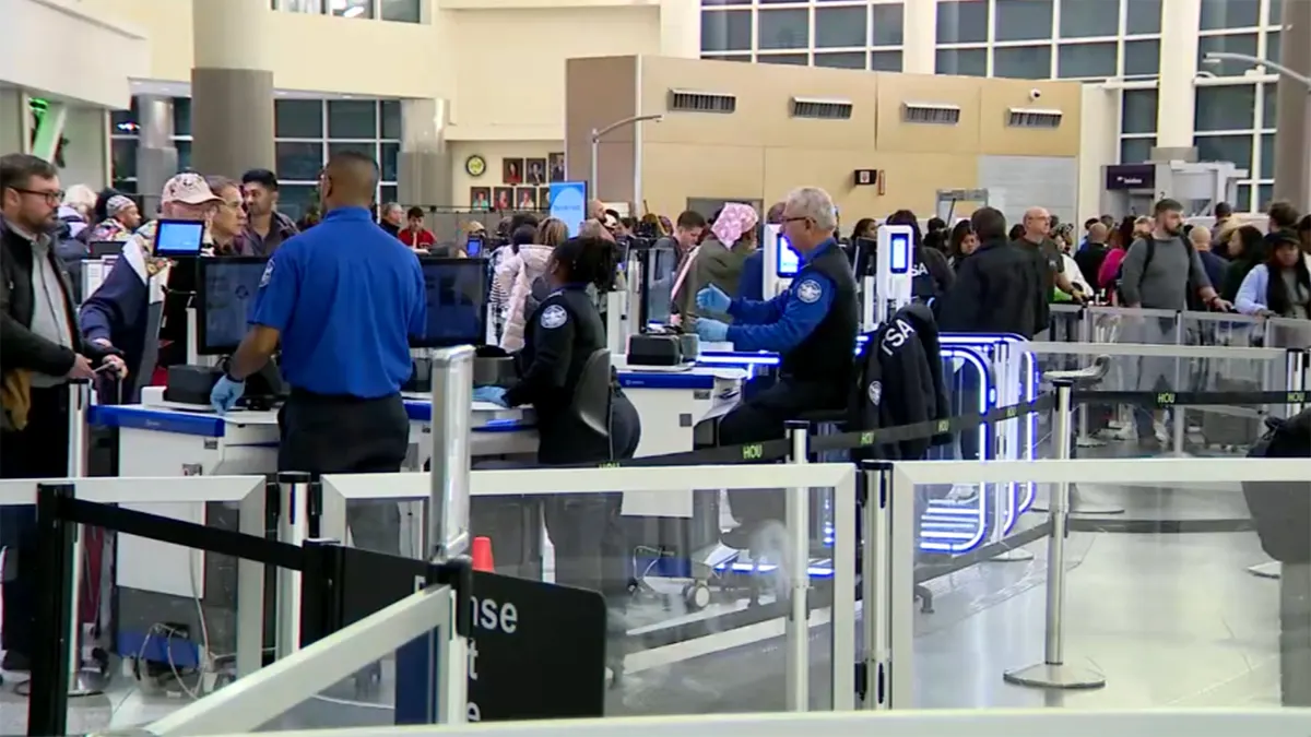 TSA agents screening passengers at airport security checkpoint with travelers in line and luggage scanners in a busy terminal.