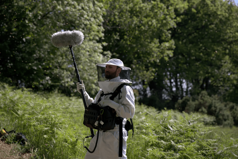 A man in a beekeeping outfit stands with a boom microphone in a field