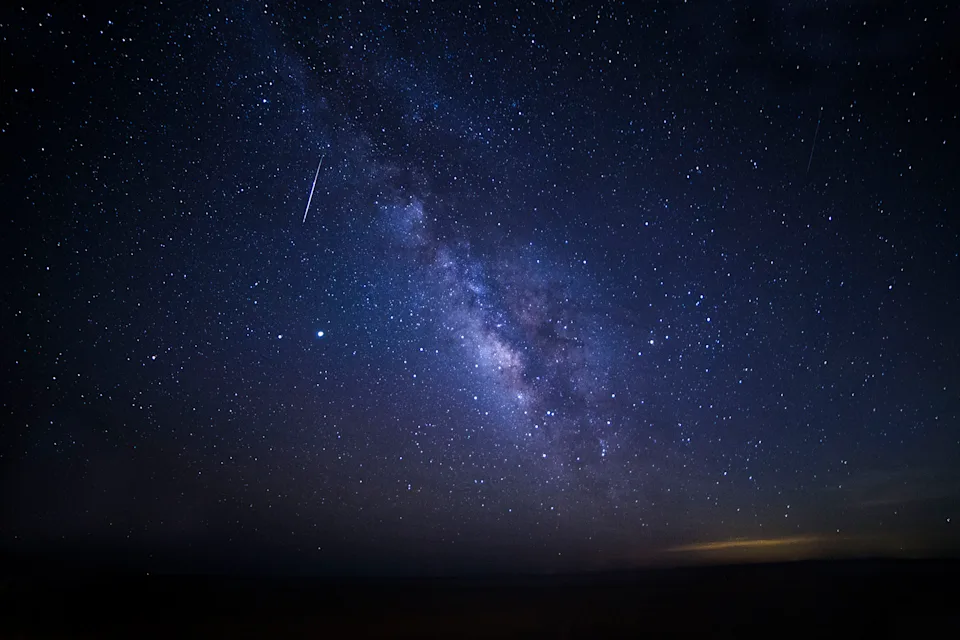 milky way streaks across the center of the image and a meteor in the top left corner leaves a long white trail in the sky. 