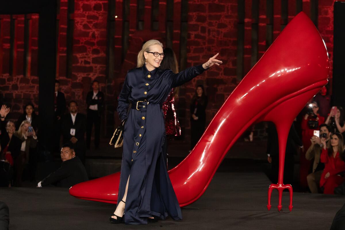 Meryl Streep smiles at the crowd while walking the carpet. There is a giant red heel behind her. 