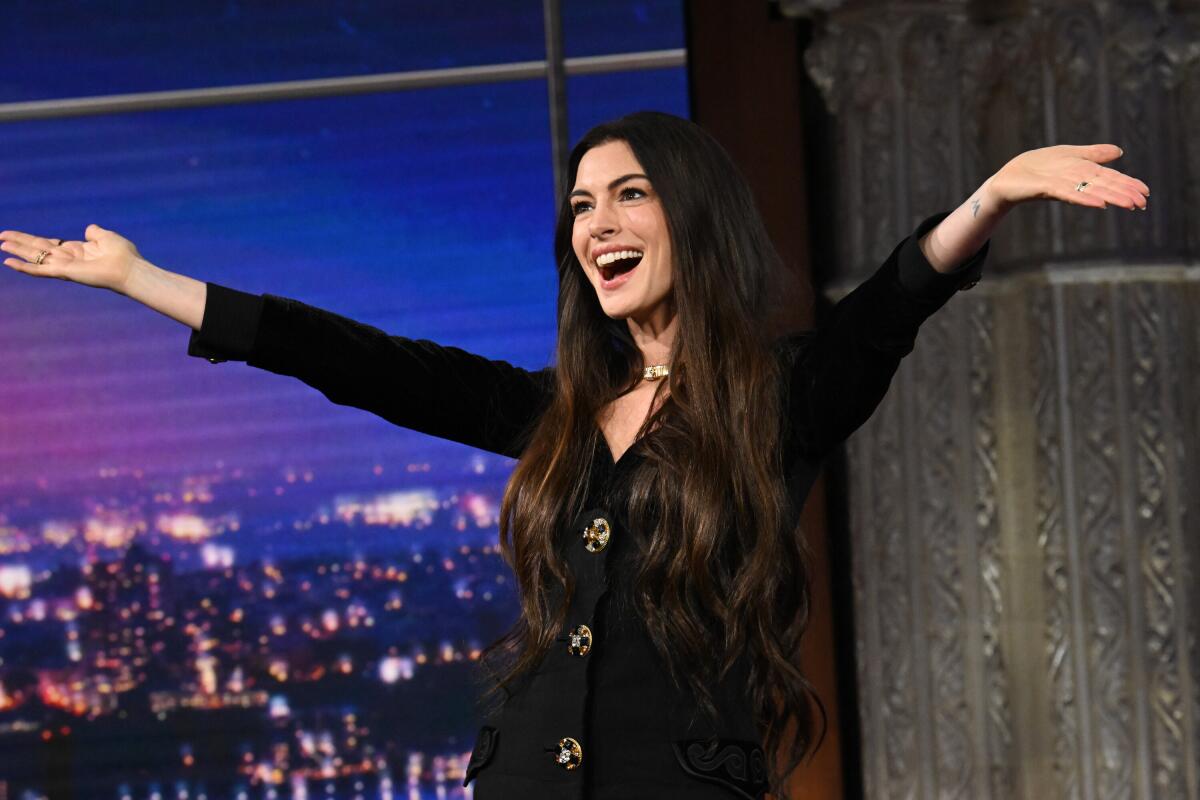 Anne Hathaway smiles with her arms up in a long-sleeved black dress.