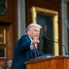 President Trump is pictured in a midrange photo at a lectern. A microphone and teleprompter are visible; he is pointing.