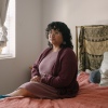 Lourdes Monje, a young person with curly dark hair and glasses, sits on a bed next to a window. They are wearing a mauve colored dress and sweater.