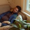 Benjamin Stein-Lobovits, a young man who has dark curly hair, a beard and glasses, lays on the couch petting his dog, Cow, a large, beige poodle mix. 