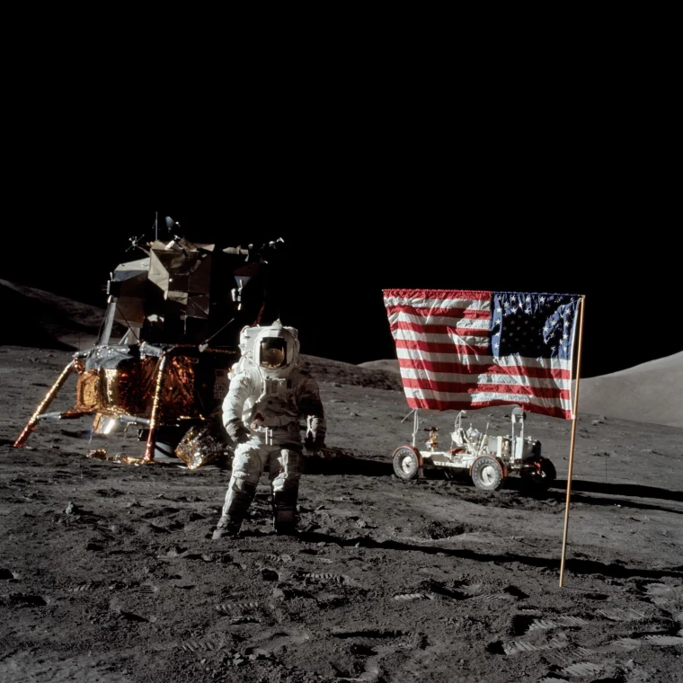 Harrison Schmitt stands on the  surface of the moon in a spacesuit, with an American flag posted in the ground on his right, and a lunar module pilot on his left