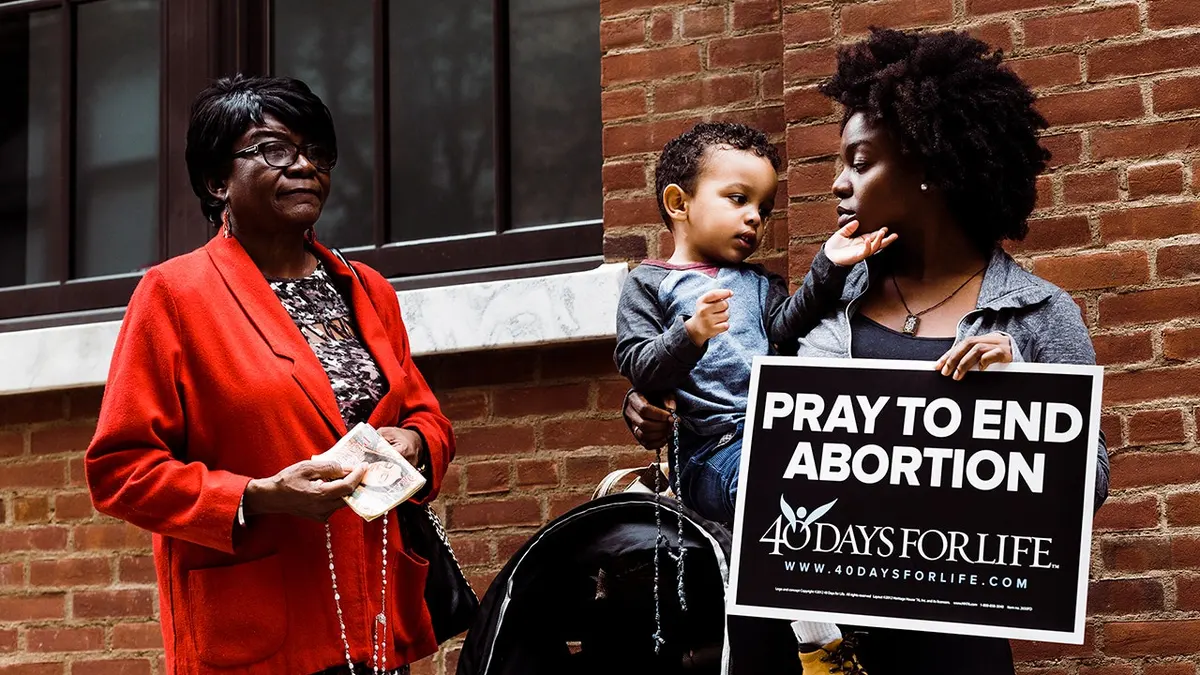 Two women and a child stand outside a building, with one holding a sign that reads 