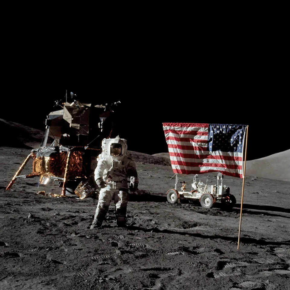 Harrison Schmitt stands on the surface of the moon in a spacesuit, with an American flag posted in the ground on his right, and a lunar module pilot on his left (JSC / NASA)