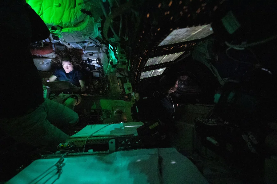 A cluttered view inside a darkened spacecraft, with a woman whose face is illuminated by a computer screen.