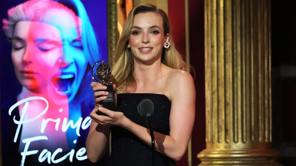 Jodie Comer pictured smiling and holding a Tony award in front of a poster for Prima Facie. She has long blonde hair and is wearing a black dress and hooped diamond earrings.