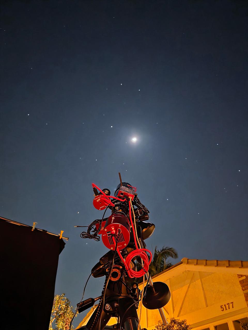 A photo of a telescope pointing at the moon in a starry night sky. The image is taken from behind the telescope and a house is visible at the bottom of the frame.