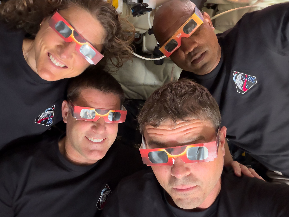 Four individuals wear red and yellow eclipse glasses. All four look at the camera.