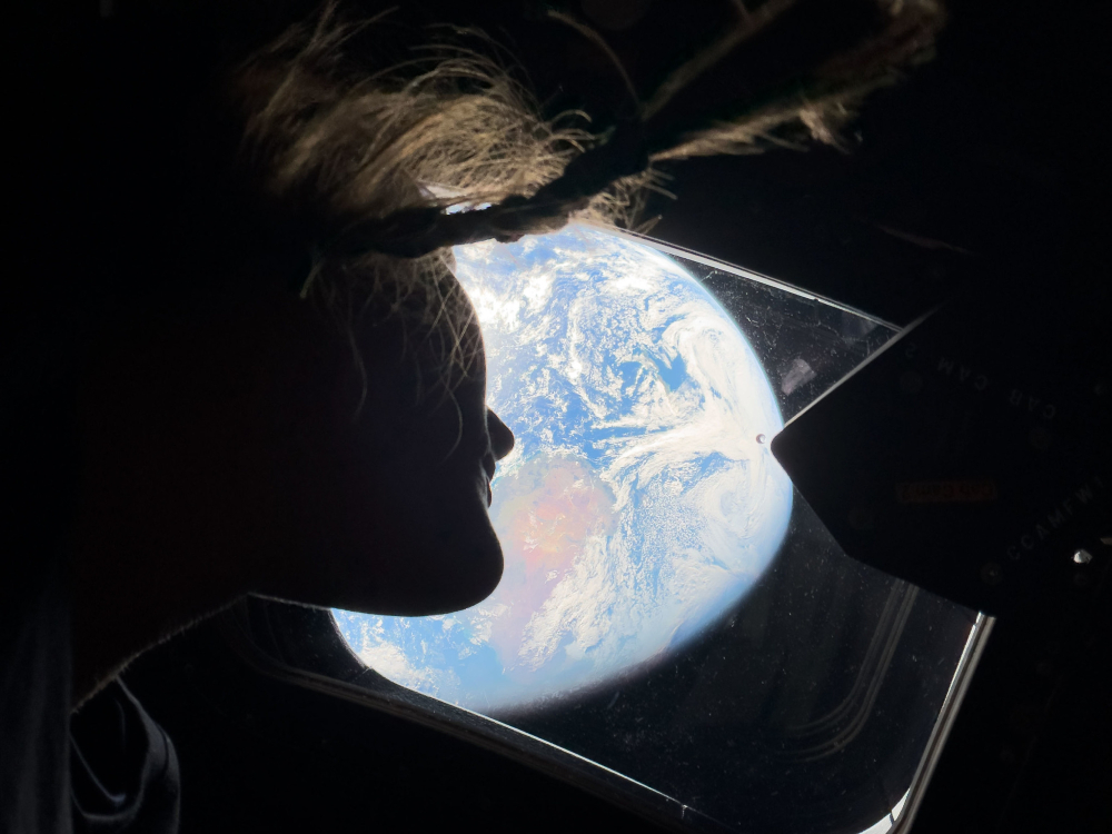 The silhouette of a woman wearing a braid is seen underneath a ceiling window showing an illuminated blue and green planet.