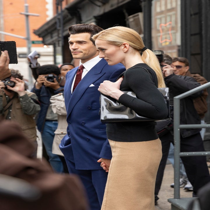 Man in a sharp suit with a red tie poses with a woman in a chic black top and beige skirt, holding a sleek bag, amid photographers