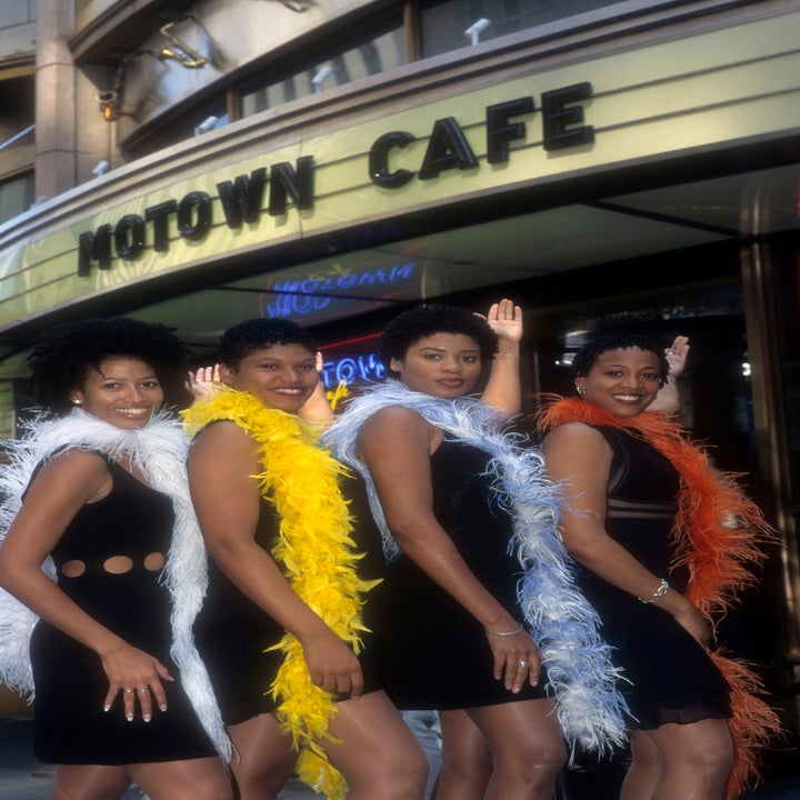 Four women wearing black dresses with feather boas pose outside Motown Cafe, raising their arms in a lively, coordinated gesture