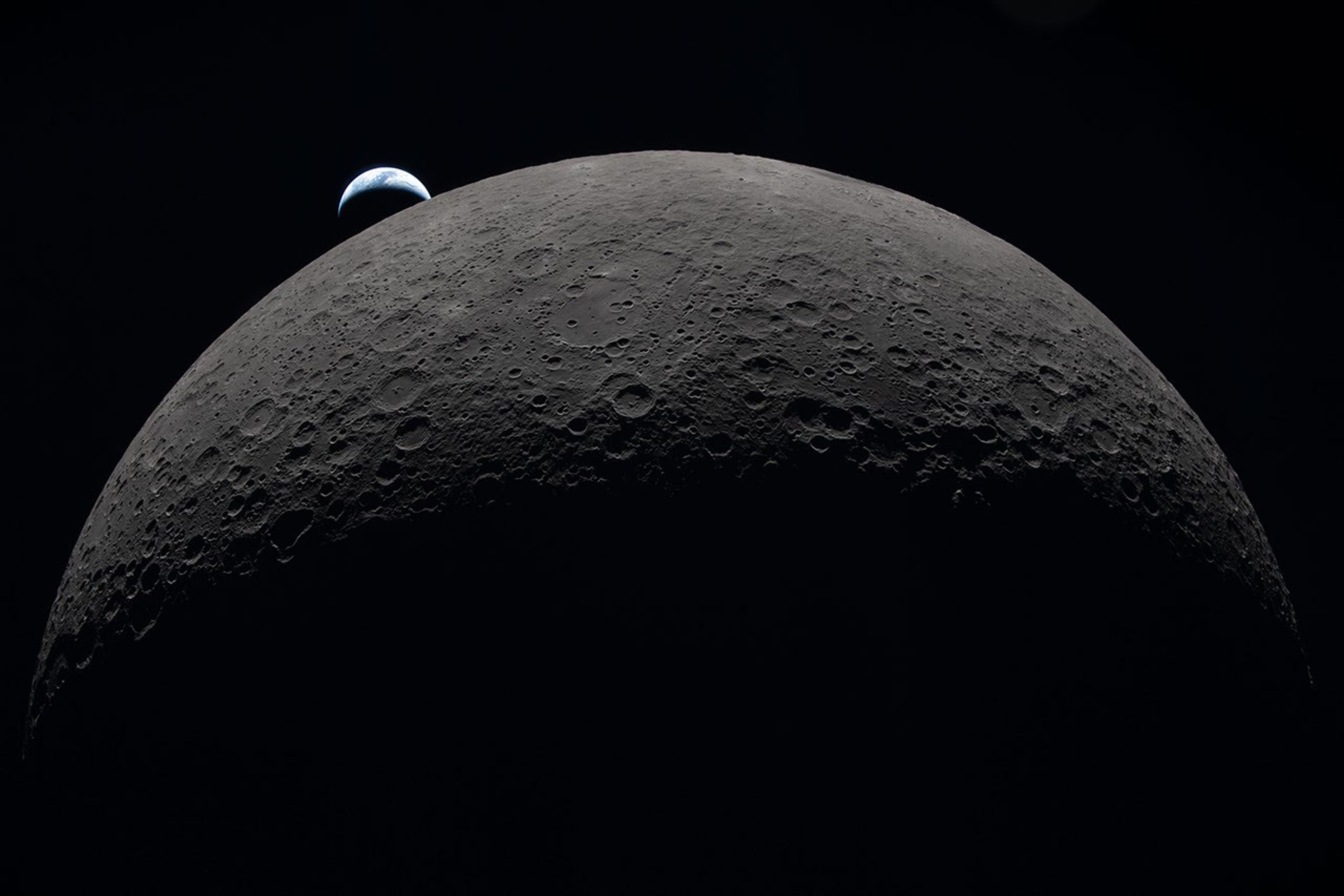 Alt text: The gray-brown, heavily cratered Moon dominates the frame against black space, with a partially lit crescent Earth setting behind its upper-left edge.