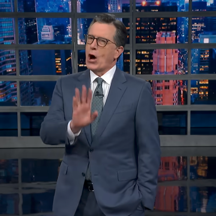 Person in a suit and glasses on a TV set speaking passionately with one hand raised