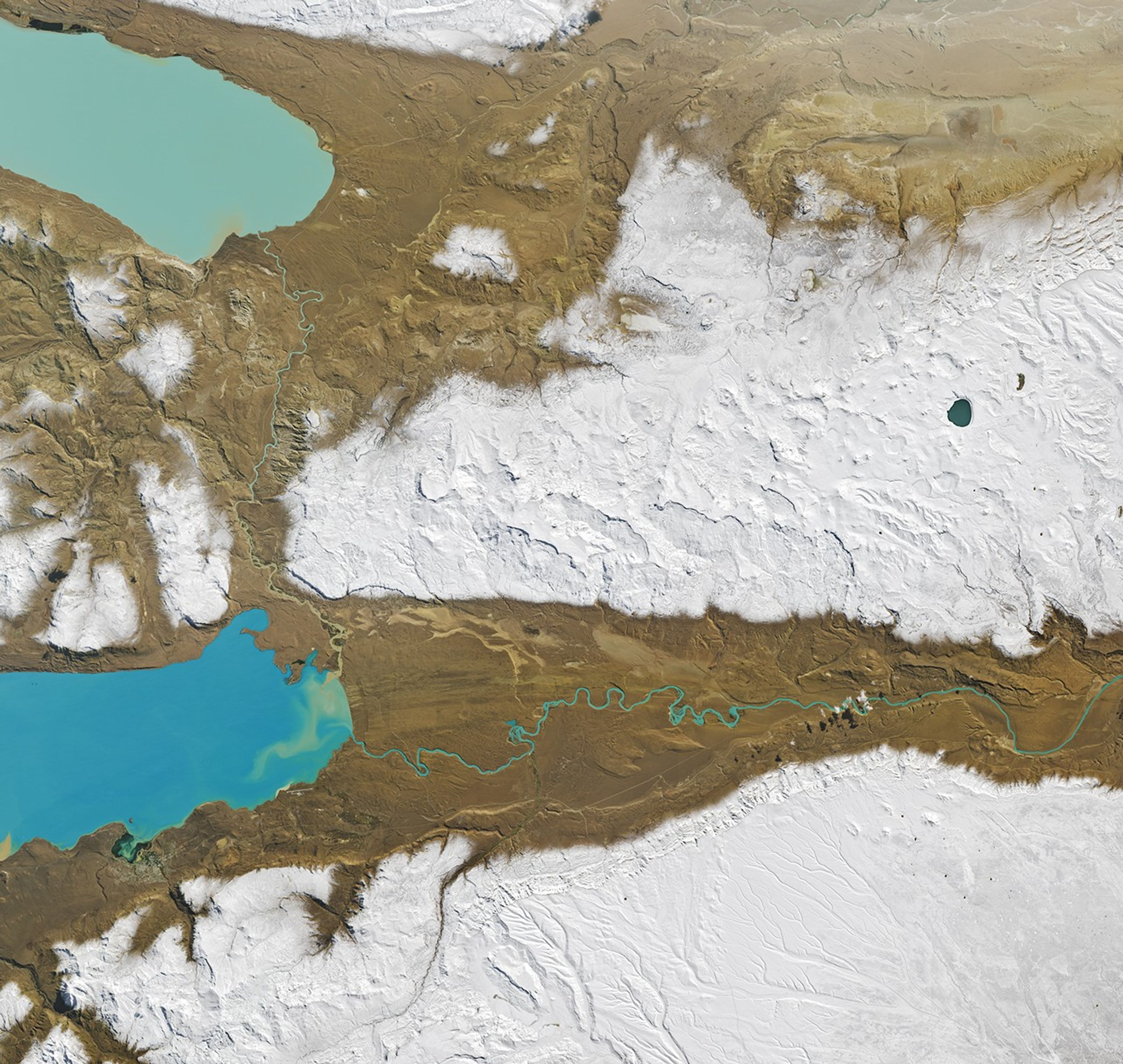 The outlet of bright blue Lago Argentino meanders across an otherwise dry, brown valley from left to right. The bottom third of the image is white, where snow covers higher elevations.