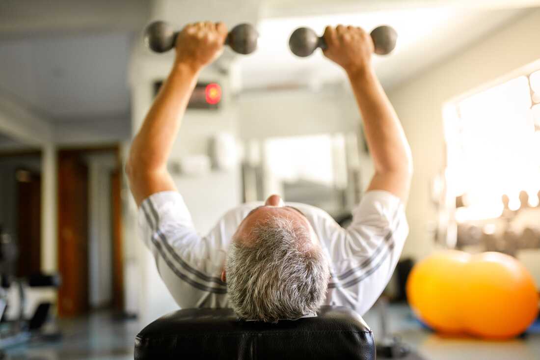 In this photo, a gray-haired man holds small dumbbells in each hand at a gym as he exercises.