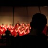 The back of a short-haired person is silhouetted in the foreground while watching a blurred choir in red robes singing in a well-lit background.