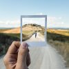 In this image, a hand holds up a Polaroid photo of a person walking down a country road in Tuscany, Italy. The photo overlaps with an actual road in Tuscany that's in view of the person holding up the photo.
