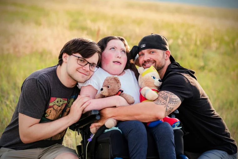 Casey Barrett and his son hug his daughter, center, who is seated in a wheelchair, she holds two teddy bears, they are all in a field