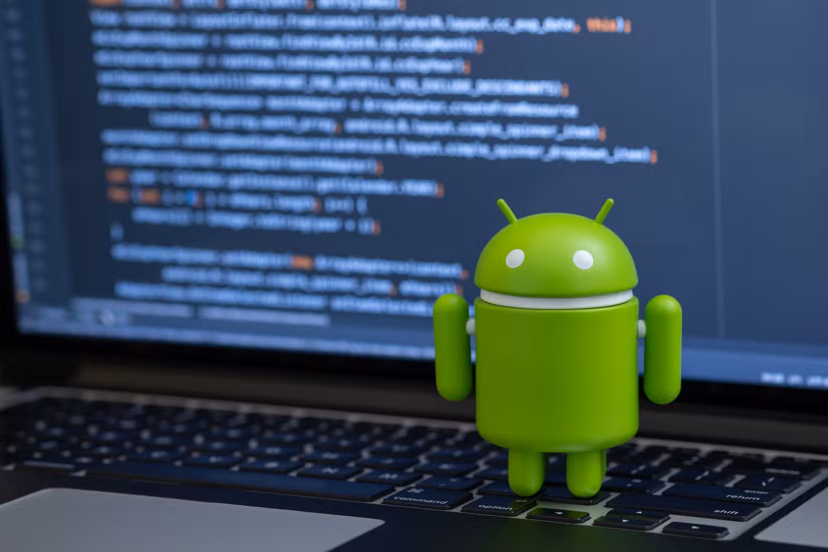 Android robot standing on a laptop keyboard with code running on the laptop screen.
