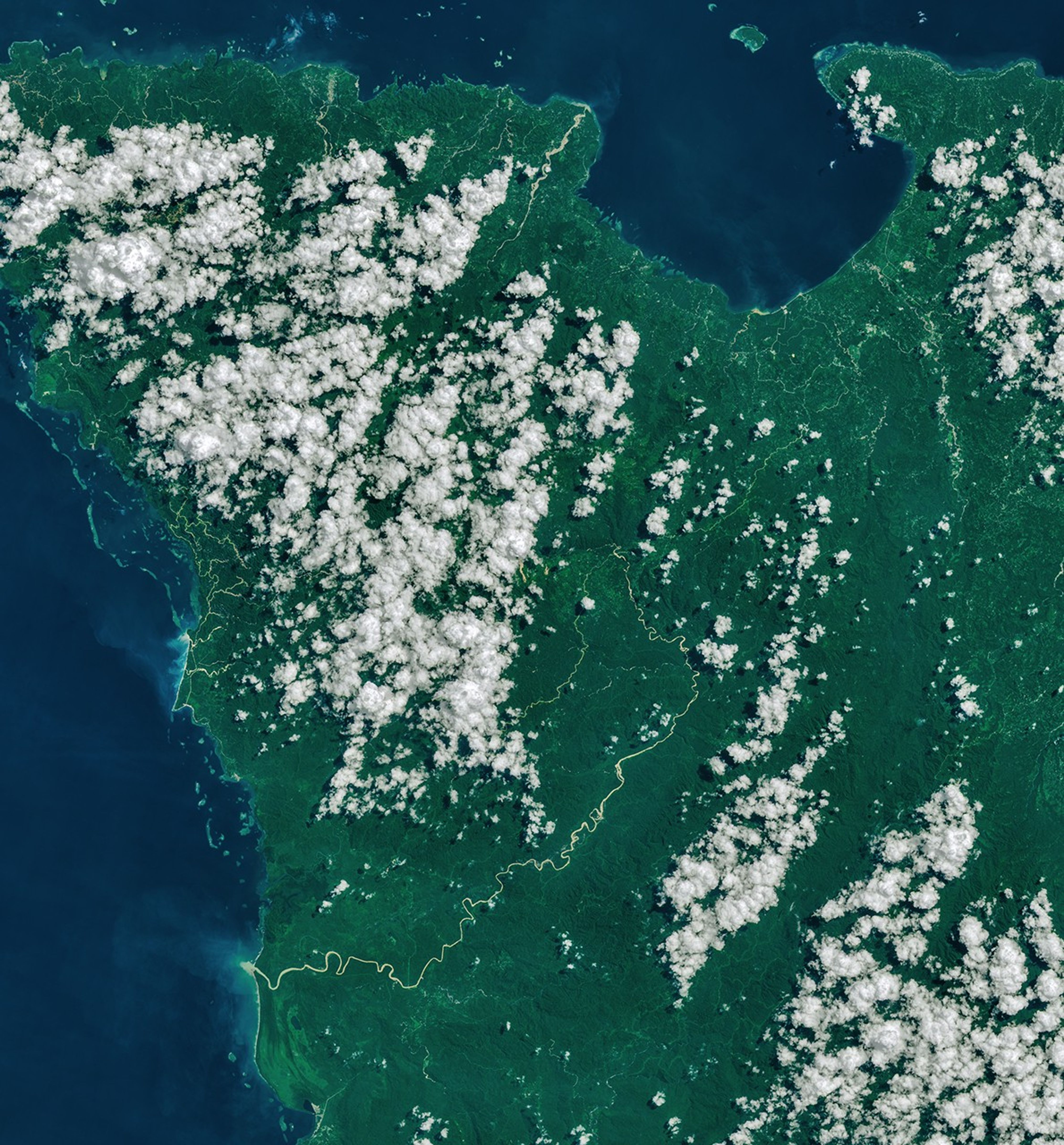 A narrow, light-brown landslide scar cuts through dark green forested mountains in East New Britain and extends south toward a stream valley. Rivers swollen with floodwaters, areas of forest clearing, and scattered clouds are also visible in some parts of the image.